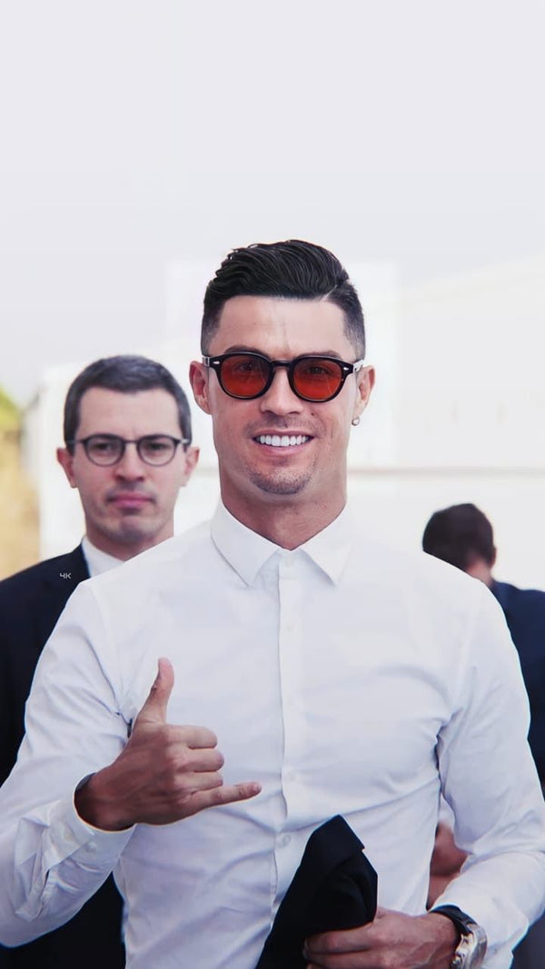 Man wearing sunglasses and a white shirt giving a thumbs-up, with another person in the background.